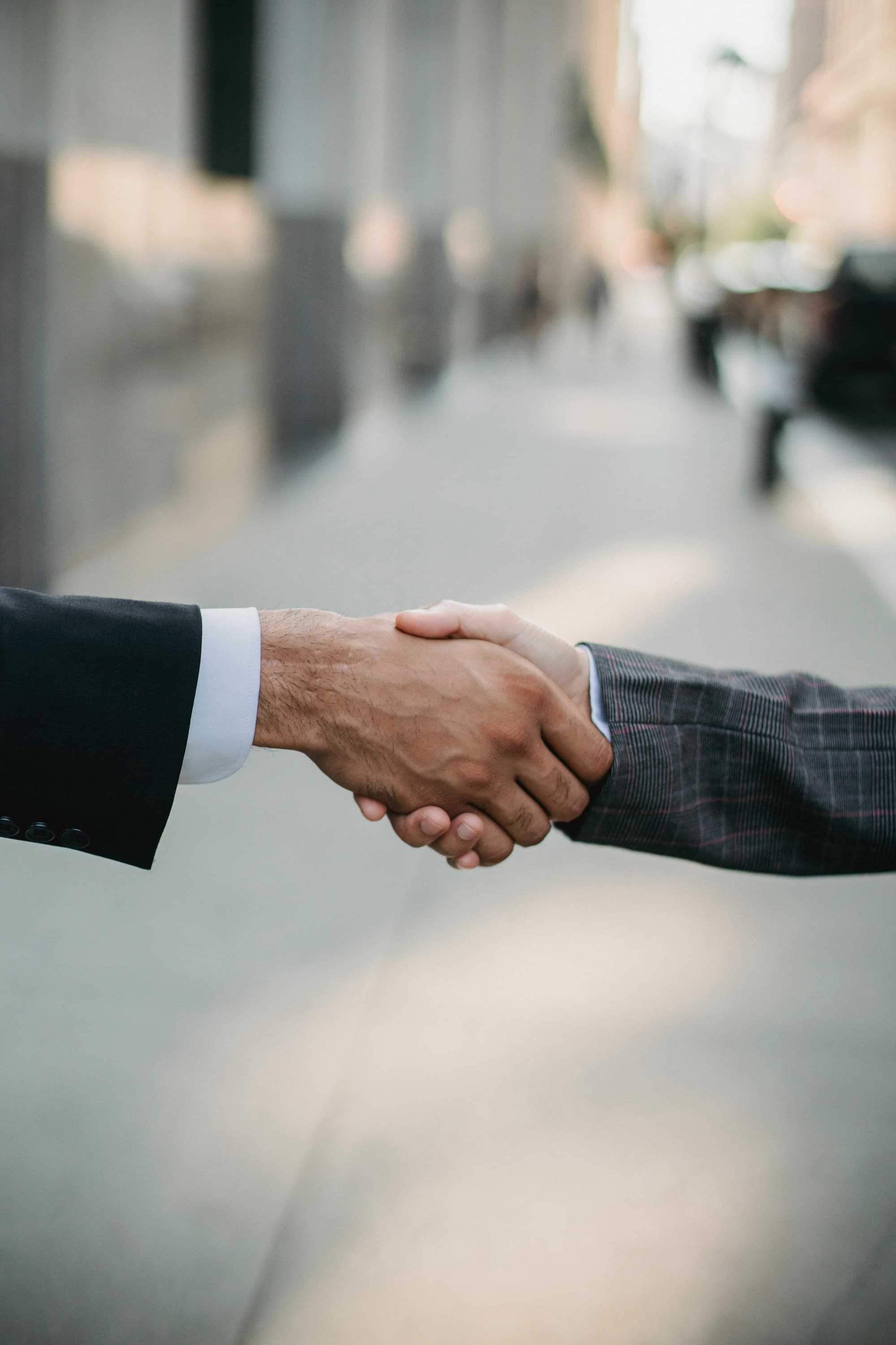 A close-up shot of two people in formal attire engaging in a handshake outdoors on a city sidewalk, with a blurred background of buildings and street activity.