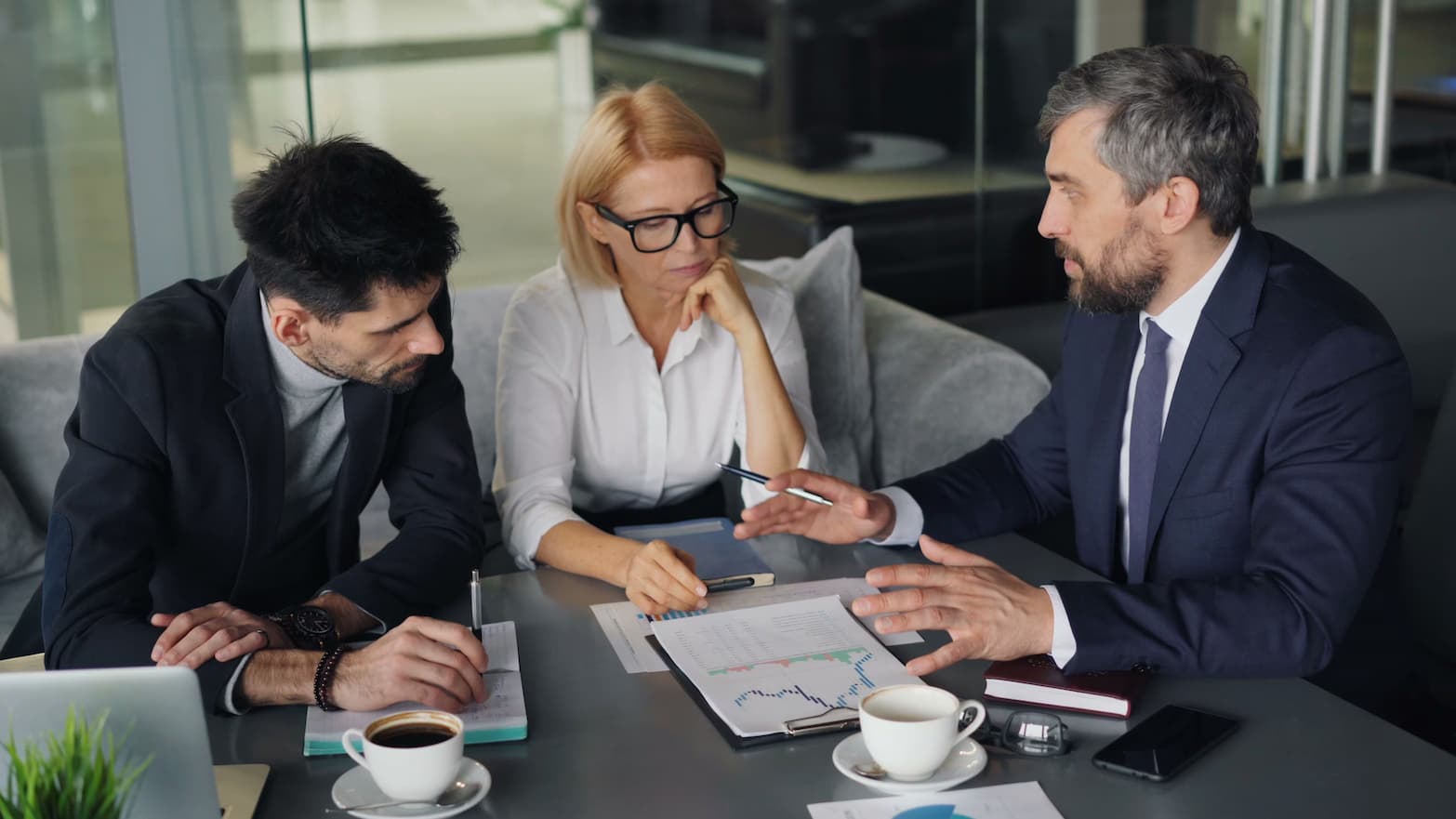 Three professionals are seated around a table in an office setting, actively discussing a financial chart laid out between them.